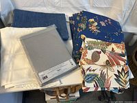 Photo of two large cloth tablecloths, multiple printed cloth napkins with floral patterns stacked and spread side by side, and a sealed pack of gray paper tablecloths.