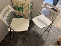 Two white IKEA folding chairs positioned on carpeted floor next to cardboard boxes.