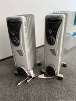 Two white Pelonis electric oil-filled radiator heaters positioned side by side on carpet. Each unit has visible power cords and caster wheels.