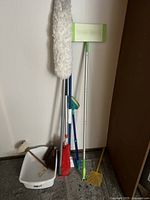Photo showing various cleaning tools including broom, dustpan, mop and duster against a white wall.