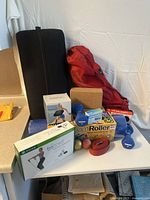The full set of yoga and fitness items displayed on a white surface with black yoga mat standing upright on the left, red bolster draped over it, cork blocks in front, blue foam roller visible, blue dumbbells in sight, pedometer and resistance band boxes visible