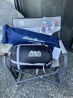 Outdoor folding camp chair with accessories on chair seat placed on gravel, showing Eddie Bauer carry bag, WFS sleeping bag, Yaktrax Pro package