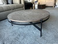 Angle view of round coffee table showing wood top and black metal frame with three legs, placed on carpet next to a couch.