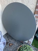 Round metal folding table folded upright against outdoor wall, gray matte finish.