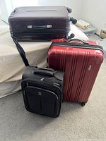 Three suitcases, two hard shell and one black soft shell, stacked near a bed on carpet.
