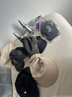 Photo showing six hats: three black baseball caps (two with 'West Seattle' embroidery and one with white 'B' logo), beige bucket hat, two metallic silver party hats, and one folded dark cloth hat.