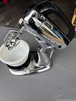Side view of Hamilton Beach electric mixer showing stainless steel body, black handle, beaters, and white milk glass bowl.