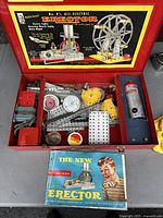 Opened metal box displaying various metal parts, electric motor, and instruction book for 1954 Erector set of a giant Ferris wheel.