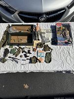 Photo showing entire lot laid out on white quilt with a Buick car in background. The lot includes a 1964 G.I. Joe figure, footlocker, manual, several uniforms, toy weapons, and the boxed John F. Kennedy G.I. Joe figure.