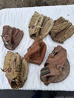 Photo showing six vintage baseball gloves arranged on a white cloth on grass, varying in color from light tan to brown, some with visible wear and damage.