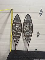 Full view of both antique snow shoes leaning against a wall with measuring tape showing height at about 42 inches.