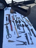 Photo showing the open antique handmade wooden toolbox with about two dozen assorted old hand tools laid out on a white cloth, including pipe wrenches, wrenches, hammers, pliers, and blow torch.