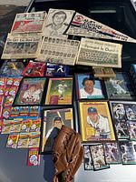 Display of newspapers, framed puzzles, baseball cards, framed oversized cards, and memorabilia including baseball gloves and pennants spread on hood of a car