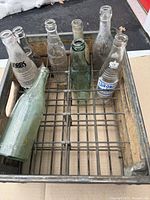 Overview of eight vintage glass soda bottles of varying brandings with wear, placed inside the wooden crate.