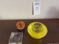 Lot of three art glass items: square clear Baccarat glass magnifier, yellow swirl Kosta Boda votive, and round floral paperweight on wood surface.