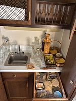 View of cabinet countertop and open drawer showing various bar ware items including glass decanters, Italian ceramic decanter figure, basket with coasters, and metal bottle openers.