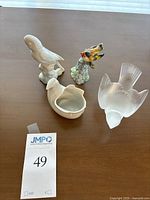 Four bird figurines on a brown table: white Dansk porcelain, colorful Cardellino bird on textured base, Lalique frosted crystal bird, and an additional white porcelain bird dish.
