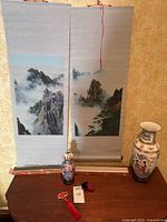 Wide view showing two foggy mountain scrolls hanging, porcelain vase on the right, cloisonné vase and decorative items on table.