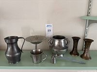 Lot showing entire collection of pewter items including pitcher, jug, compote, cup, carved vessels, measuring cup and tool on shelf against beige wall with auction number sign