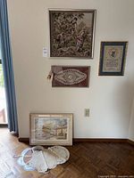 Wide angle view of lot including framed needlepoint tapestry, crochet doily, printed art, dinner roll holders on floor, and framed text needlepoint