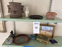 Overview of two shelves showing large terracotta planter, glass sand vial, decorative dishes, small sculptures, painted boxes, framed painting.