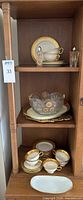 Wooden cabinet shelf holding gold and white Lenox China pieces including plates, tea cups with saucers, and serving bowls, plus cut glass serving pieces and a pair of salt and pepper shakers