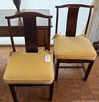 Front view of two identical Asiatic rosewood chairs with yellow cushions tied on seat cushions, placed side by side on a wooden floor with a side table behind.