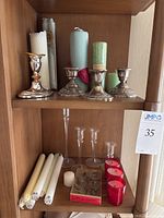 Shelf with three sterling silver weighted candlesticks and assorted beeswax candles behind them, plus white taper candles, small white candle and red votive candles below on another shelf