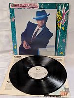 Photo of vinyl LP record with white label and track information on the turntable alongside the album cover featuring Elton John in a blue suit and hat
