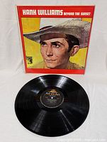 Photo showing front cover of Hank Williams Beyond The Sunset LP with portrait of Hank Williams in cowboy hat, colorful background, and MGM label visible on vinyl disc.