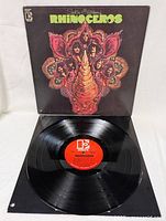 Vinyl record with red center label showing album and artist information lying in front of the album cover which features psychedelic rhinoceros artwork.