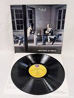 Vinyl record with yellow label and album cover showing two mannequins sitting at a table in a sparse room.