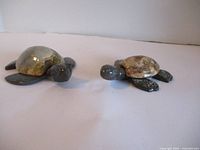 Photo showing two marble turtle figurines positioned facing each other on a flat white surface, highlighting their polished stone shells and detailed carvings on heads and limbs.