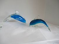 Two art glass dolphin figurines on a white surface, showing size comparison and clear with blue coloring