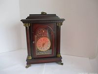 Front view of the wooden Bombay mantle clock showing the clock face, columns, and decorative brass details.
