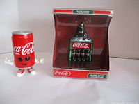 Two Coca-Cola themed ornaments displayed together: a character can figure standing beside a boxed glass bottle carrier ornament.