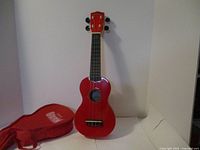 Front view of the red Mahalo soprano ukulele highlighting the body, fingerboard and tuning pegs, placed beside the red soft case