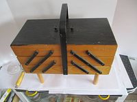 Closed view of the wooden sewing box with black metal handles and side supports, showing the exterior design and legs.