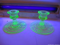 Photo under UV light showing both uranium glass candle holders glowing green, raised etched floral pattern visible on bases.