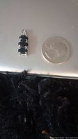 Close-up photo of the sterling silver sapphire pendant shown next to a quarter for size comparison. The pendant has three dark blue stones in a vertical row.
