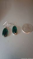 Pair of vintage sterling silver earrings with oval translucent green helenite stones set in silver hooks, placed next to a US quarter coin to show size.