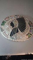 Close-up photo showing the silver-toned oval belt buckle with Native American chief silhouette in black inlay at the center, surrounded by abalone shell, turquoise, and coral inlay. Detailed engraved patterns on metal surface visible.