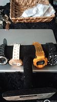 Four watches on a black display stand including Geneva rhinestone quartz watch with black strap, Geneva rhinestone bezel watch with white strap, yellow digital Smirnoff Orange Twist watch, and black Always kids watch.