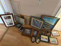 Wide view of collection of framed paintings and empty picture frames on the floor against wall, showing variety of artwork and frames.