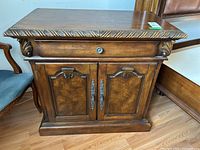 Front view showing the carved decorative accents on the top edge and ornate metal handles on the cabinet doors. Visible scratches and scuffs on wood surface.