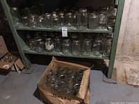 Wide view of two shelf levels holding multiple clear glass canning jars with metal lids and a box on the floor with more jars.