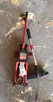 Overall view of the red Motomaster hydraulic trolley jack with attached handle and the car brush laying beside it on the floor.