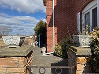 Wide outdoor image showing both matching square concrete planters positioned on stone pedestals beside a brick building under daylight sky.
