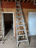 Full view of the aluminum 10-foot stepladder standing upright against a plywood wall in a garage or workshop environment, showing the entire ladder and its step structure.