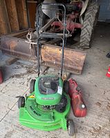 Green Lawn Boy push lawn mower with Dura Force engine and two red jerry cans on concrete floor in garage with rusty equipment in background.